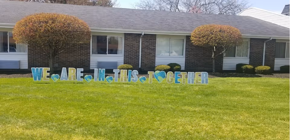 A green lawn in front of a brick building with two small trees and trimmed bushes. Large colorful letters on the grass spell out 'WE ARE IN THIS TOGETHER' with blue heart shapes between some words.