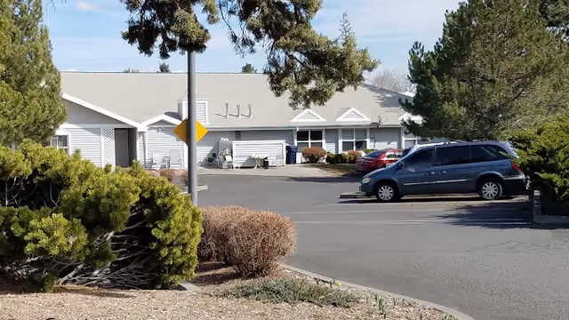 Exterior view of a single-story building with a light gray roof and white siding, surrounded by trees and bushes. Several cars are parked in the parking lot in front of the building under a clear sky.