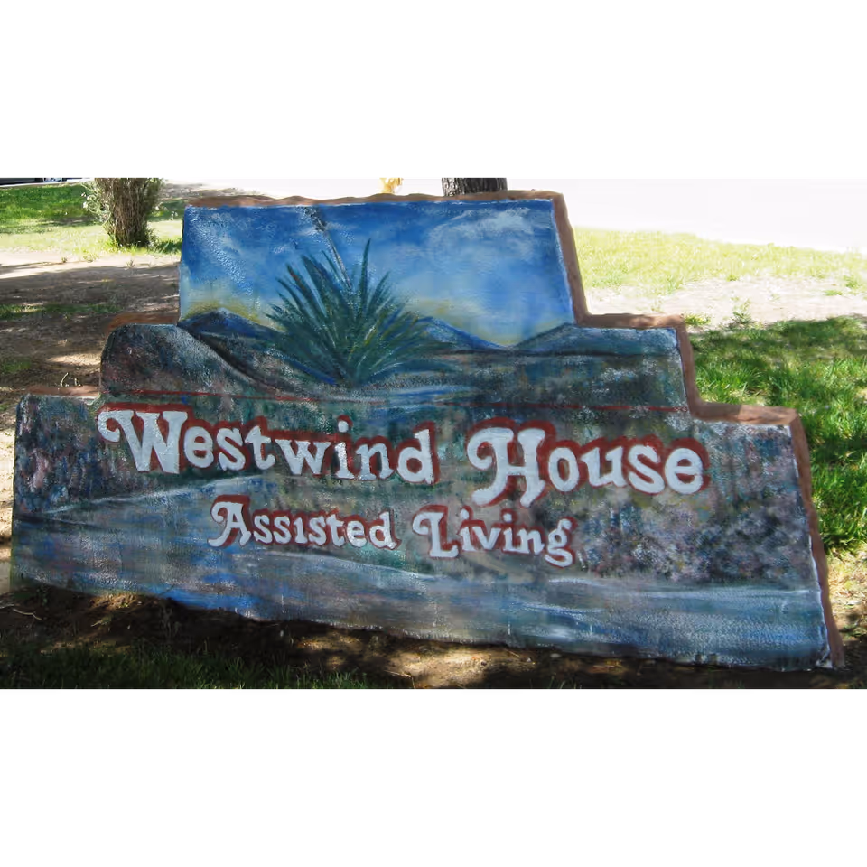 A painted stone sign outdoors with the text 'Westwind House Assisted Living' surrounded by grass and trees.