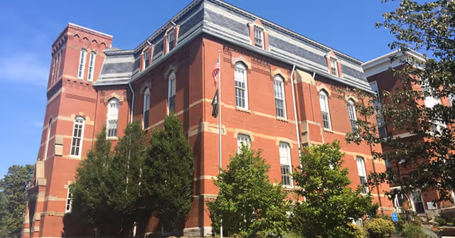 Red-brick multi-story building with flags and trees in front under a blue sky.