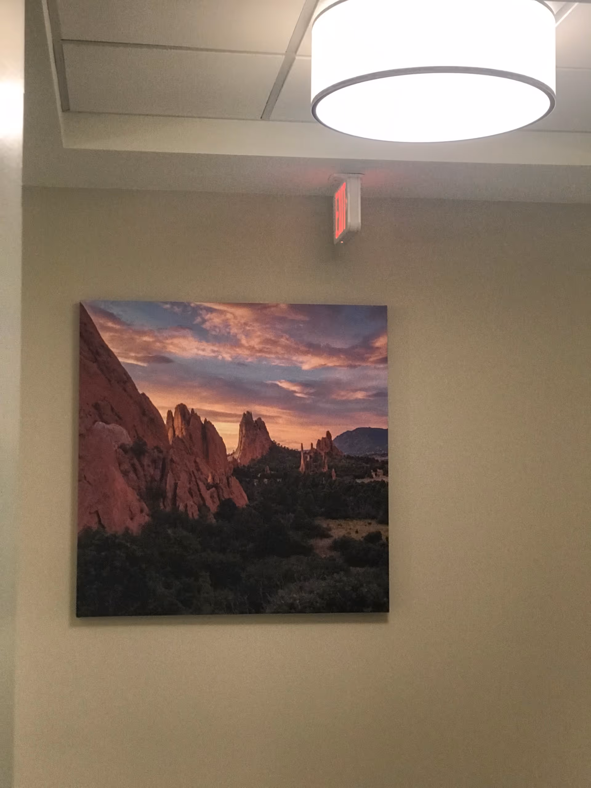 A wall inside a building with a large landscape photograph of rocky formations and a colorful sunset sky. Above the photograph is a round ceiling light fixture and an exit sign is visible near the ceiling.
