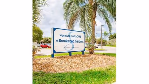 White sign reading 'Signature HealthCARE of Brookwood Gardens' set in mulch bed with palm trees and a street in the background.
