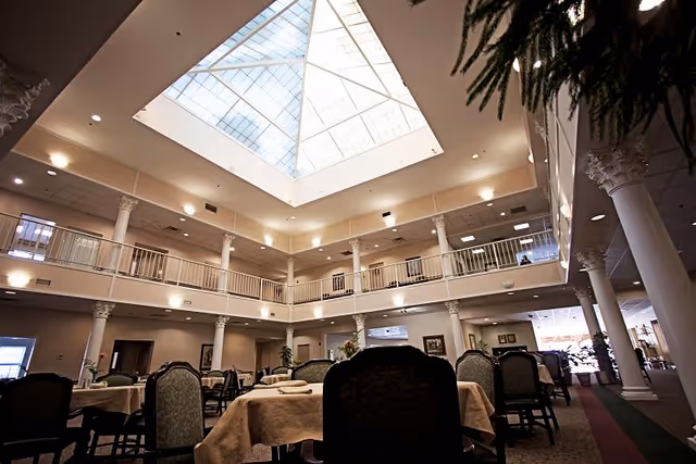 Interior view of a spacious dining area in a senior living facility with tables covered in beige tablecloths and chairs arranged around them. The room features tall white columns, a second-floor balcony railing, and a large skylight ceiling allowing natural light to fill the space.
