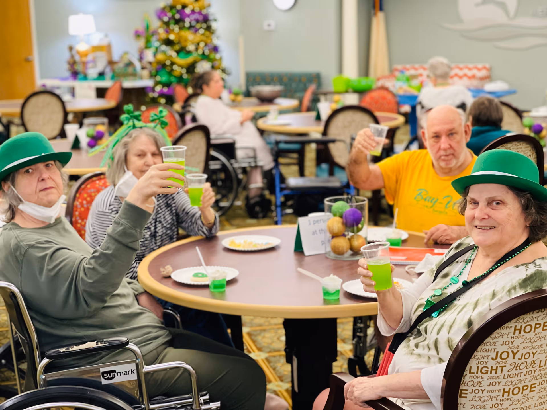 A group of elderly people sitting around a round table in a decorated room, raising cups with green drinks in a toast. Two women wear green hats and festive accessories, and there is a Christmas tree in the background. Plates with snacks and a decorative centerpiece are on the table.