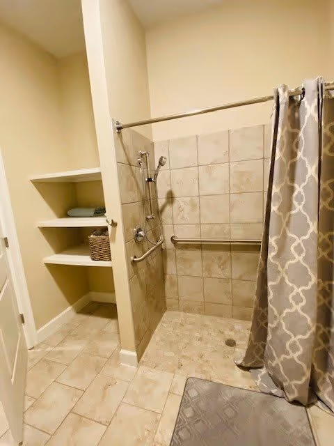 A bathroom shower area with beige tiled walls and floor, equipped with a handheld showerhead and a horizontal grab bar. A patterned gray and white shower curtain is partially drawn to the side. To the left, there are built-in shelves holding folded towels and a wicker basket. The bathroom has light-colored walls and a tiled floor.