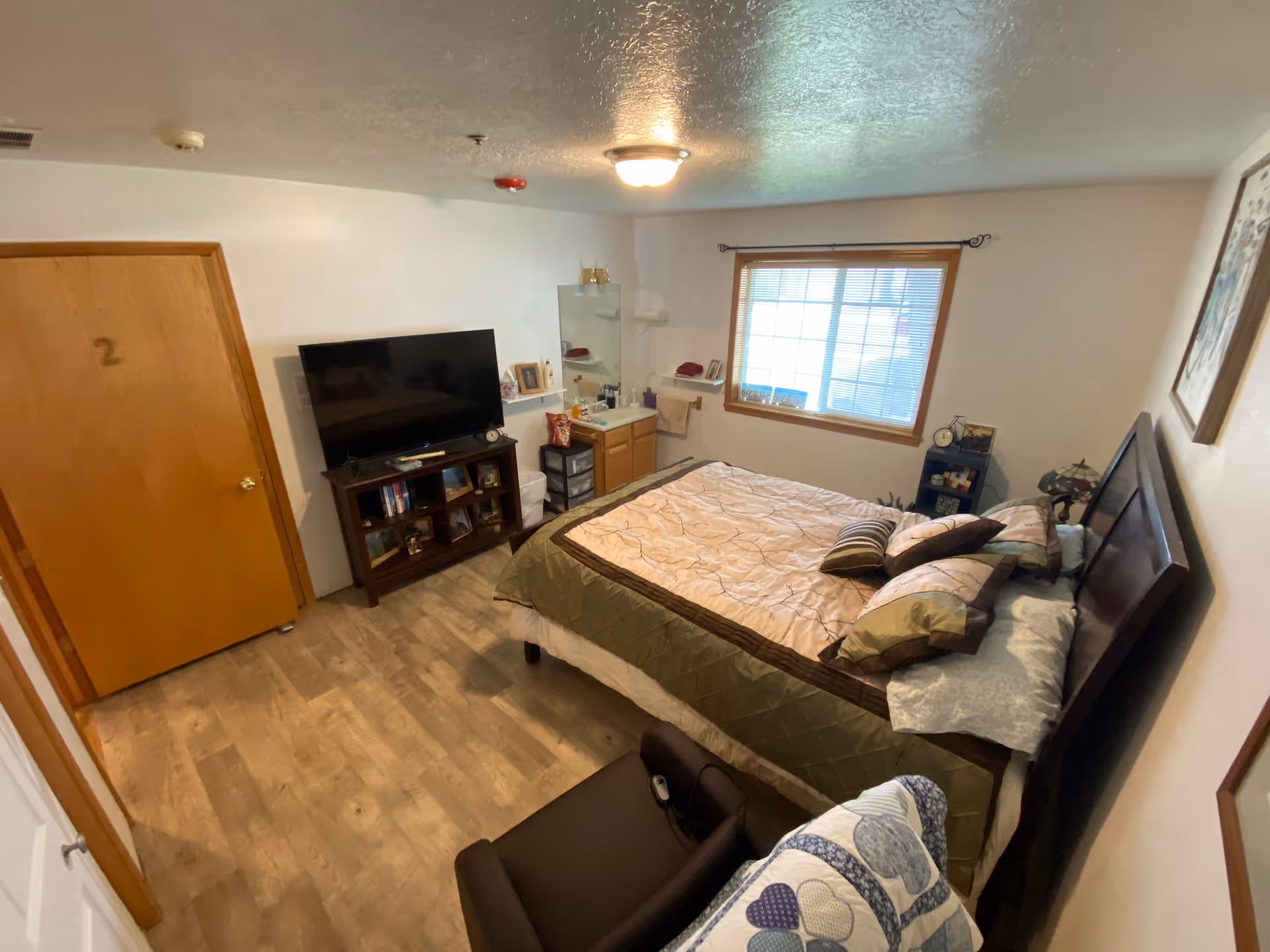 A cozy bedroom in an assisted living facility with a bed covered in a beige and brown quilt, several pillows, a window with blinds, a wooden door marked with the number 2, a TV on a wooden stand with shelves, a small sink with a mirror, and a cushioned chair with a patterned blanket draped over it.