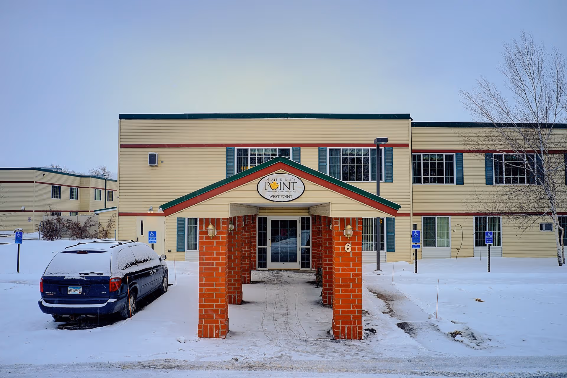 Exterior view of Nature's Point Assisted Living building in winter with snow covering the ground and a blue vehicle parked in a handicapped parking spot near the entrance. The building is two stories tall with beige siding, green window shutters, and red trim. A covered walkway with red brick pillars leads to the main entrance.