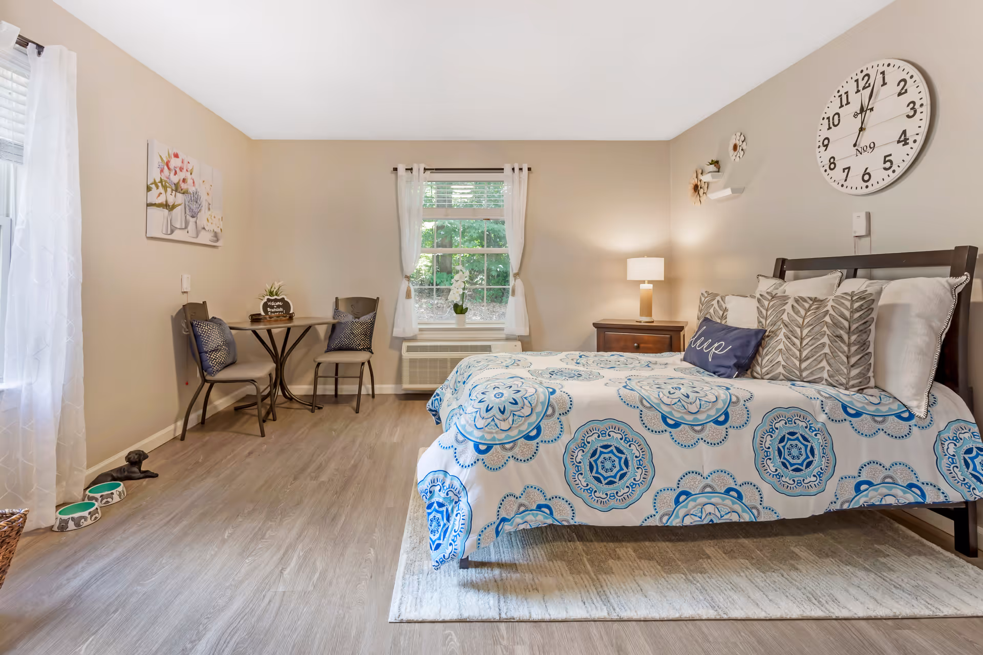 A cozy bedroom with a bed featuring a blue and white patterned bedspread and multiple pillows, including one with the word 'sleep'. There is a wooden nightstand with a lamp beside the bed. A round wall clock hangs above the bed. A small table with two chairs is placed near a window with white curtains, and there is a wall painting of flowers. The floor is wooden with a light-colored rug under the bed. Two pet bowls are on the floor near the window.