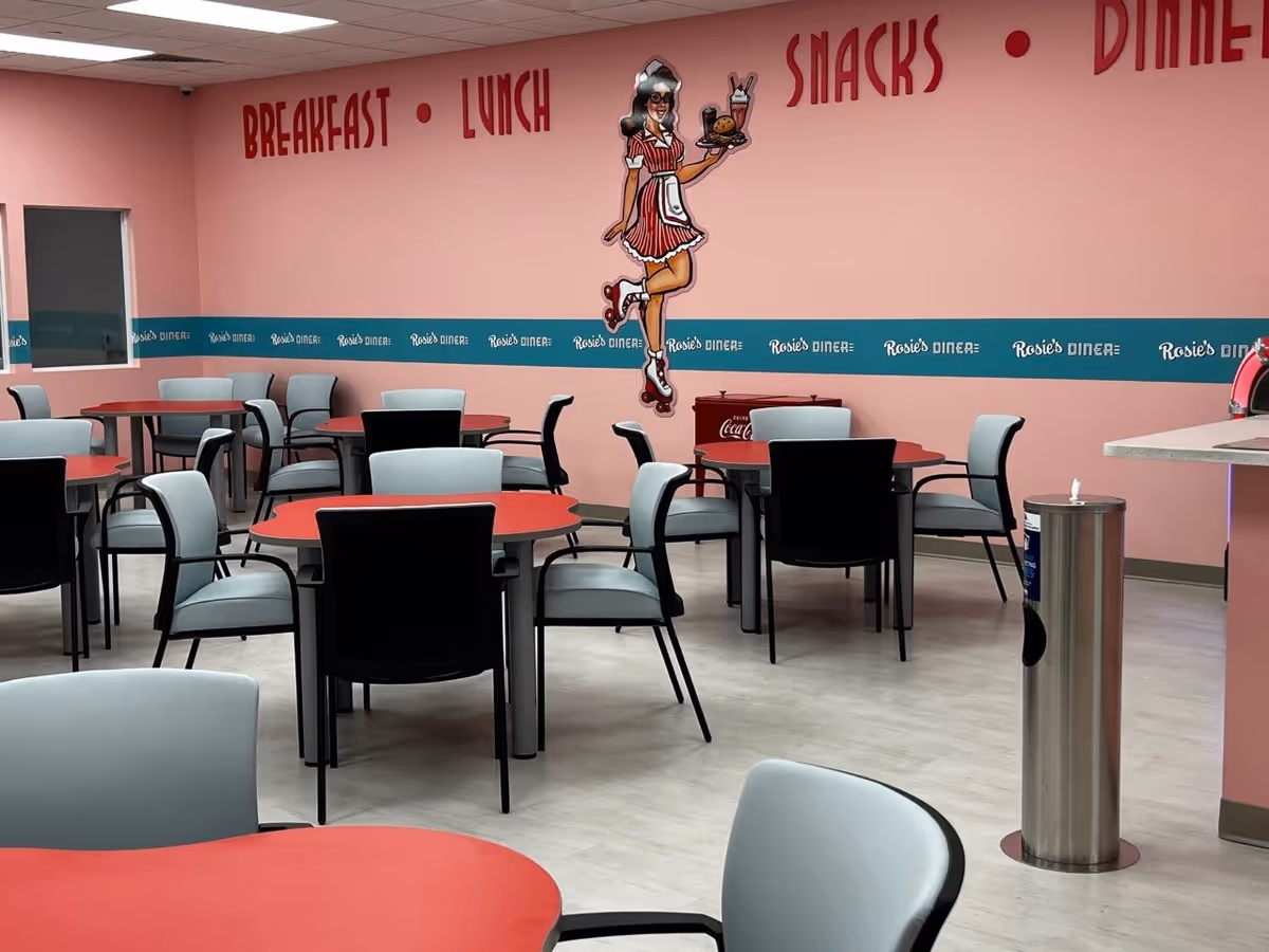 Interior view of a dining area with red and black tables and gray chairs. The walls are painted pink with a teal stripe that reads 'Rosie's Diner' repeatedly. Above the stripe, large red letters spell out 'BREAKFAST', 'LUNCH', 'SNACKS', and 'DINNER'. A retro-style illustration of a waitress holding a tray with a burger and a milkshake is displayed on the wall. The floor is light-colored, and there is a stainless steel trash can near the counter on the right.