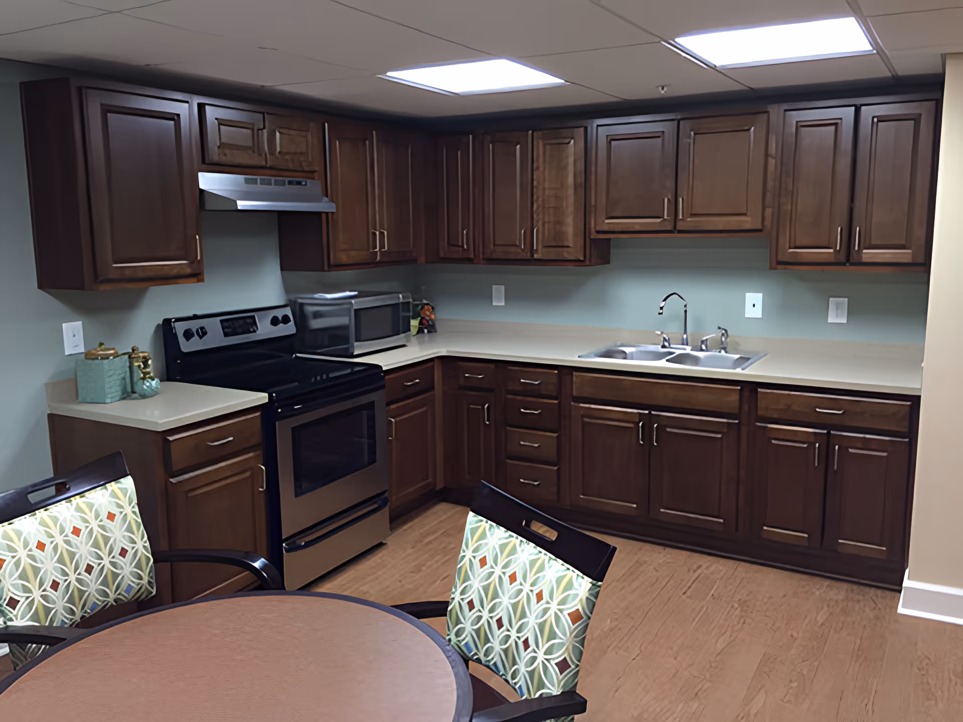 A kitchen area with dark wooden cabinets, a stainless steel stove with an overhead vent, a microwave on the countertop, a double sink, and a round table with chairs that have patterned cushions.