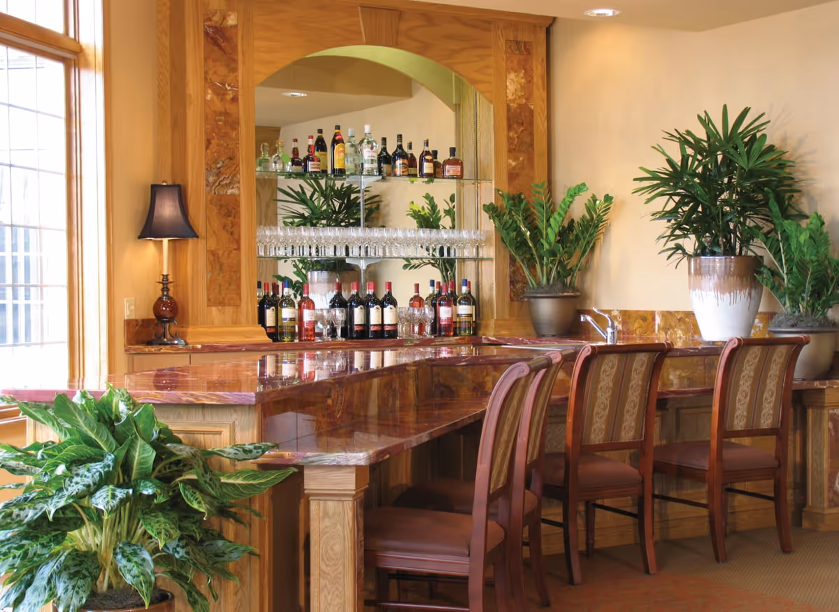 Interior view of a senior living facility bar area with a polished wooden counter, four cushioned wooden chairs, several potted green plants, a table lamp, and shelves stocked with various bottles of wine and liquor along with glassware.