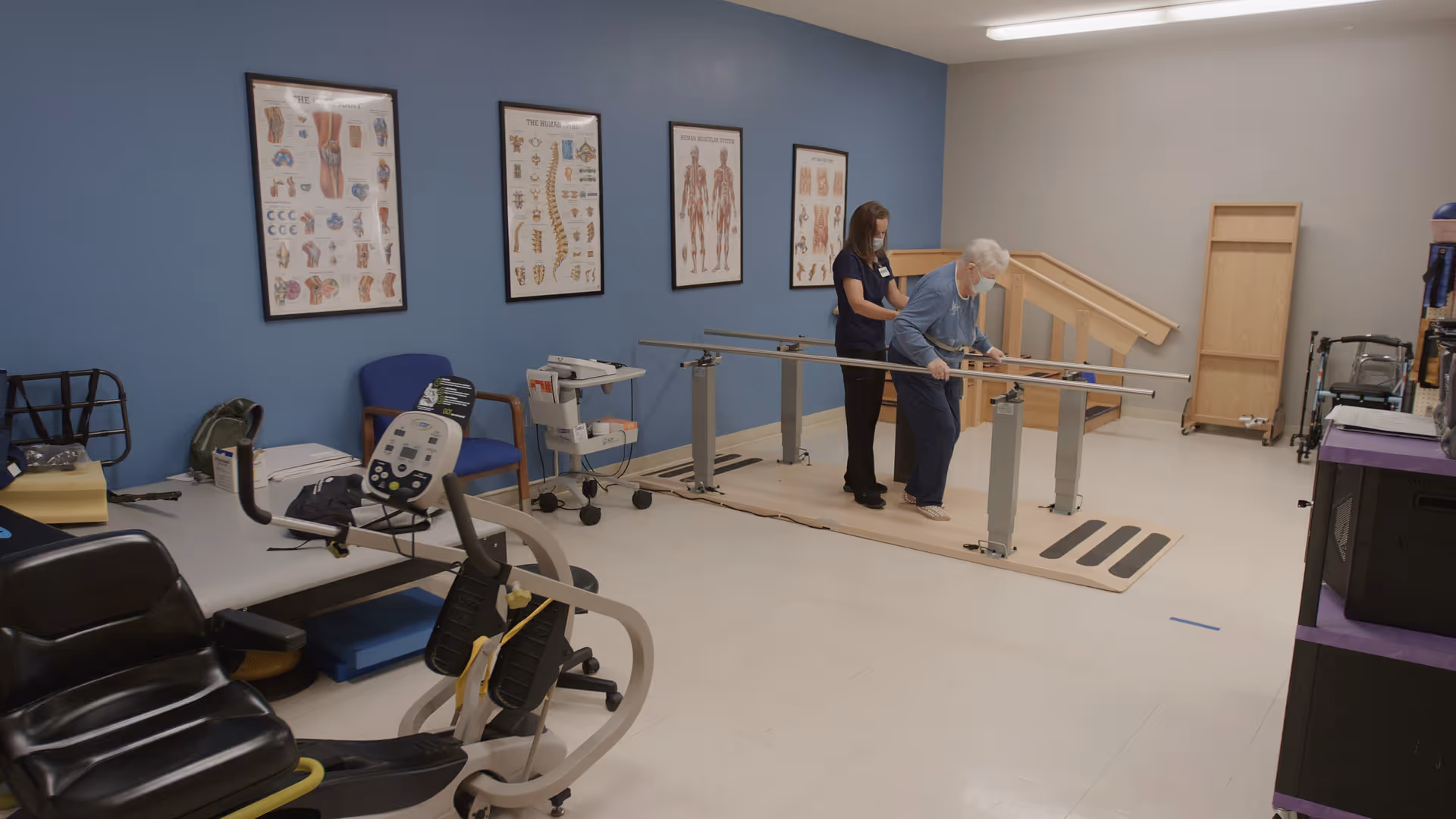 A physical therapy room with an elderly woman using parallel bars for walking practice, assisted by a healthcare worker wearing a mask. The room has exercise equipment, anatomical posters on a blue wall, and a small set of stairs with a handrail in the background.