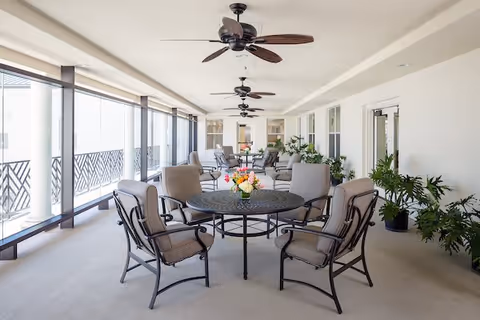 A spacious, covered outdoor patio area with multiple round tables surrounded by cushioned chairs. Ceiling fans are mounted overhead, and potted plants line the walls. Large windows with decorative railings allow natural light to fill the space.