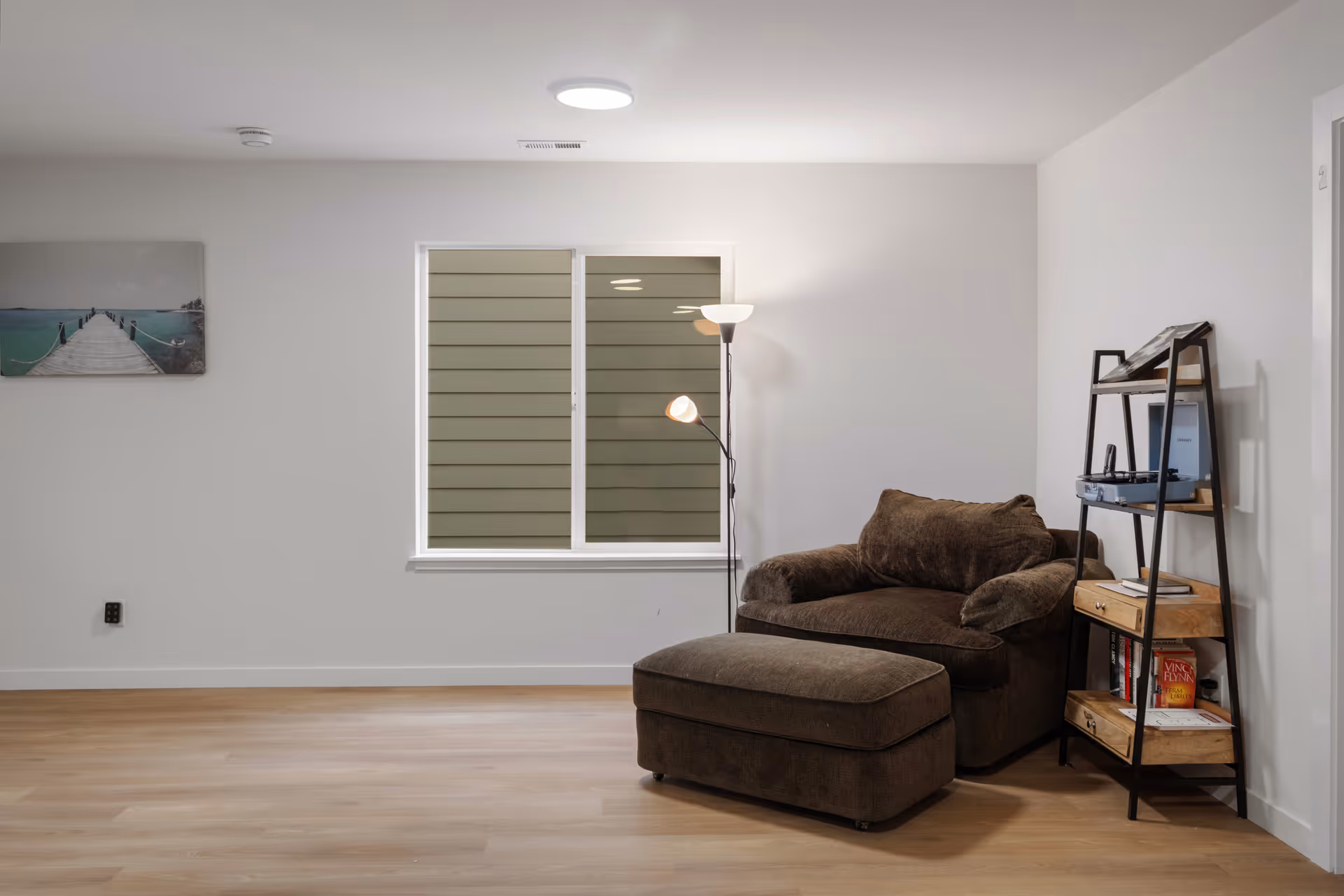 A cozy living room corner with a large brown armchair and matching ottoman. Next to the chair is a black metal and wood shelving unit holding books and a record player. A floor lamp with two lights stands behind the chair. A window with closed blinds is centered on the wall, and a framed picture of a pier over water hangs on the left wall. The room has light wood flooring and white walls.
