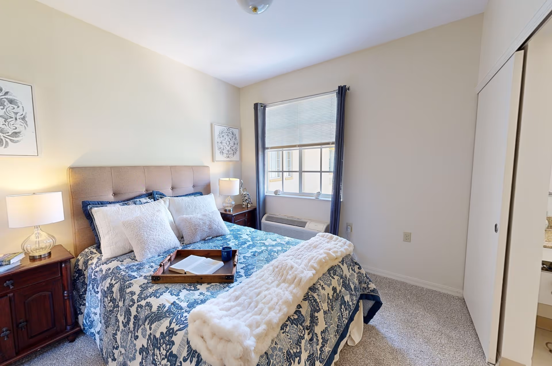A cozy bedroom with a bed featuring a tufted beige headboard, blue and white floral bedding, and multiple pillows. There is a wooden tray on the bed holding an open book and a blue mug. Two bedside tables with lamps flank the bed. A window with blinds and dark curtains is on the wall behind the bed, and a white closet door is visible on the right side. The room has light-colored walls and carpeted flooring.