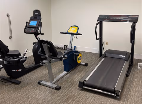 A small exercise room with three pieces of fitness equipment: a recumbent stationary bike, an upright stationary bike, and a treadmill. The room has beige walls and carpeted flooring.