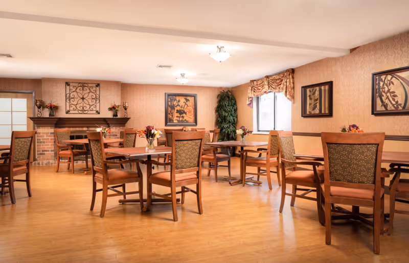 A warm and inviting dining room with wooden tables and chairs arranged neatly. The room features a brick fireplace with decorative items on the mantle, framed artwork on the walls, a large potted plant near a window with patterned curtains, and small floral centerpieces on each table. The floor is wooden, and the lighting is soft and ambient.
