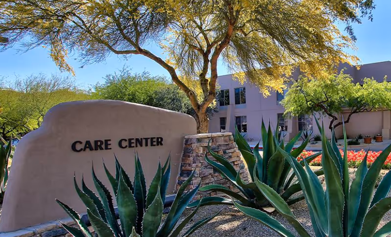 Outdoor view of a care center building with desert landscaping including agave plants and trees, under a clear blue sky.