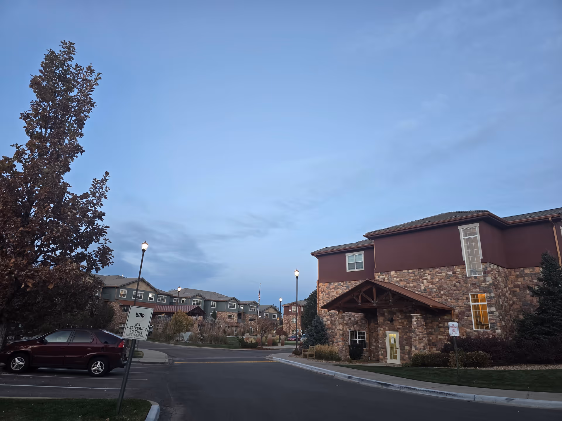 Exterior front of a brick-and-stucco senior living building with parking, lamp posts, and landscaped grounds under a clear evening sky.