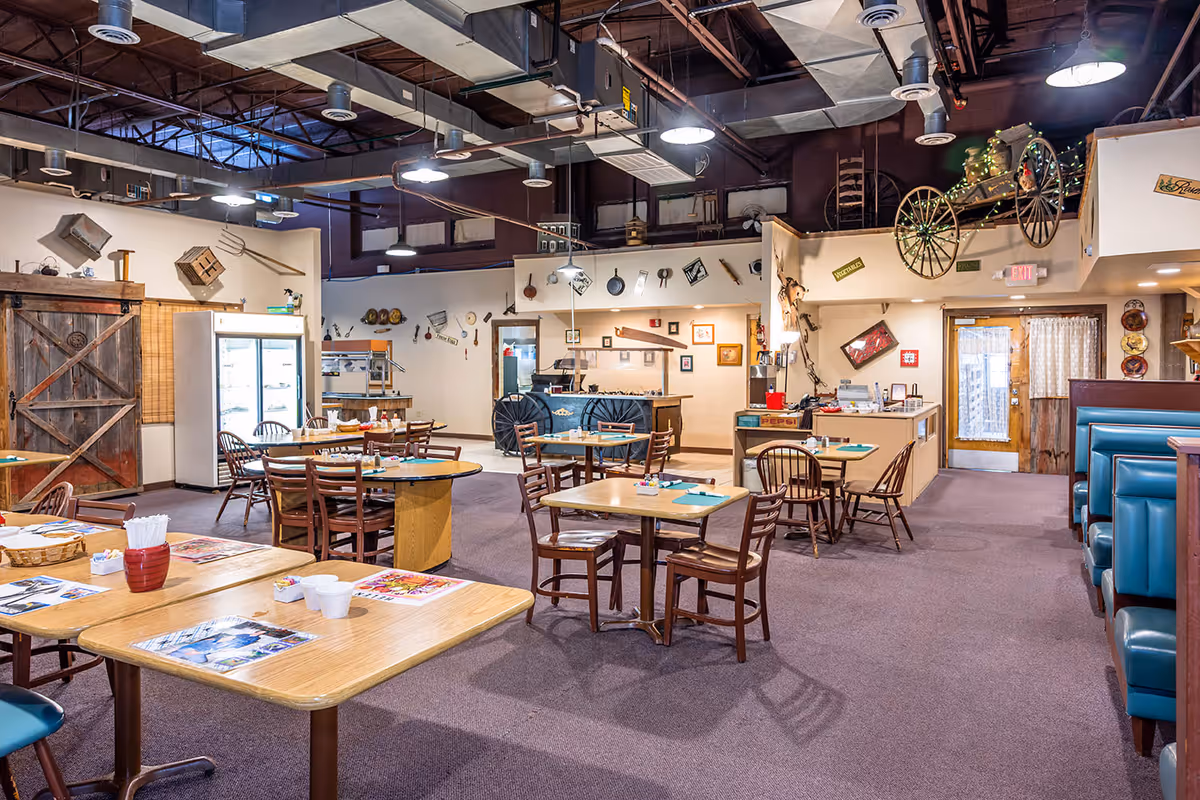 Interior view of a dining area with wooden tables and chairs, teal booth seating, rustic decor including a wooden barn door, antique farming tools on the walls, and a wagon wheel mounted above. The space has an industrial ceiling with exposed ducts and lighting fixtures.
