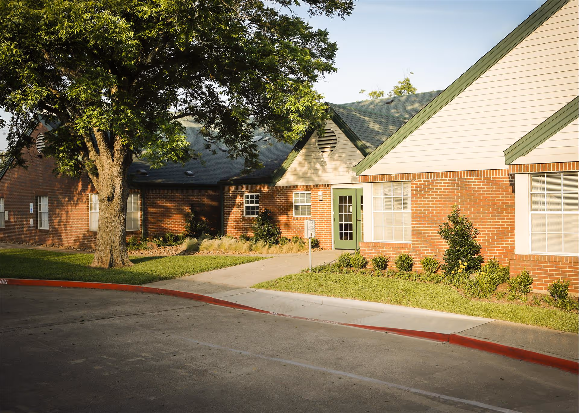 Exterior view of a brick building with green-trimmed roof and doors, surrounded by a well-maintained lawn, shrubs, and a large tree, under a clear sky.