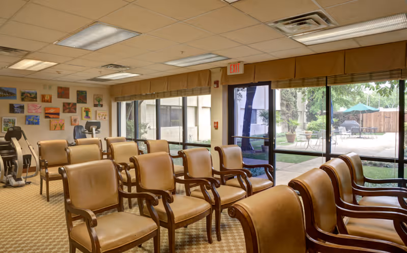 Sunlit common room with rows of upholstered chairs facing a wall of windows overlooking a patio, with exercise equipment and artwork on the walls.