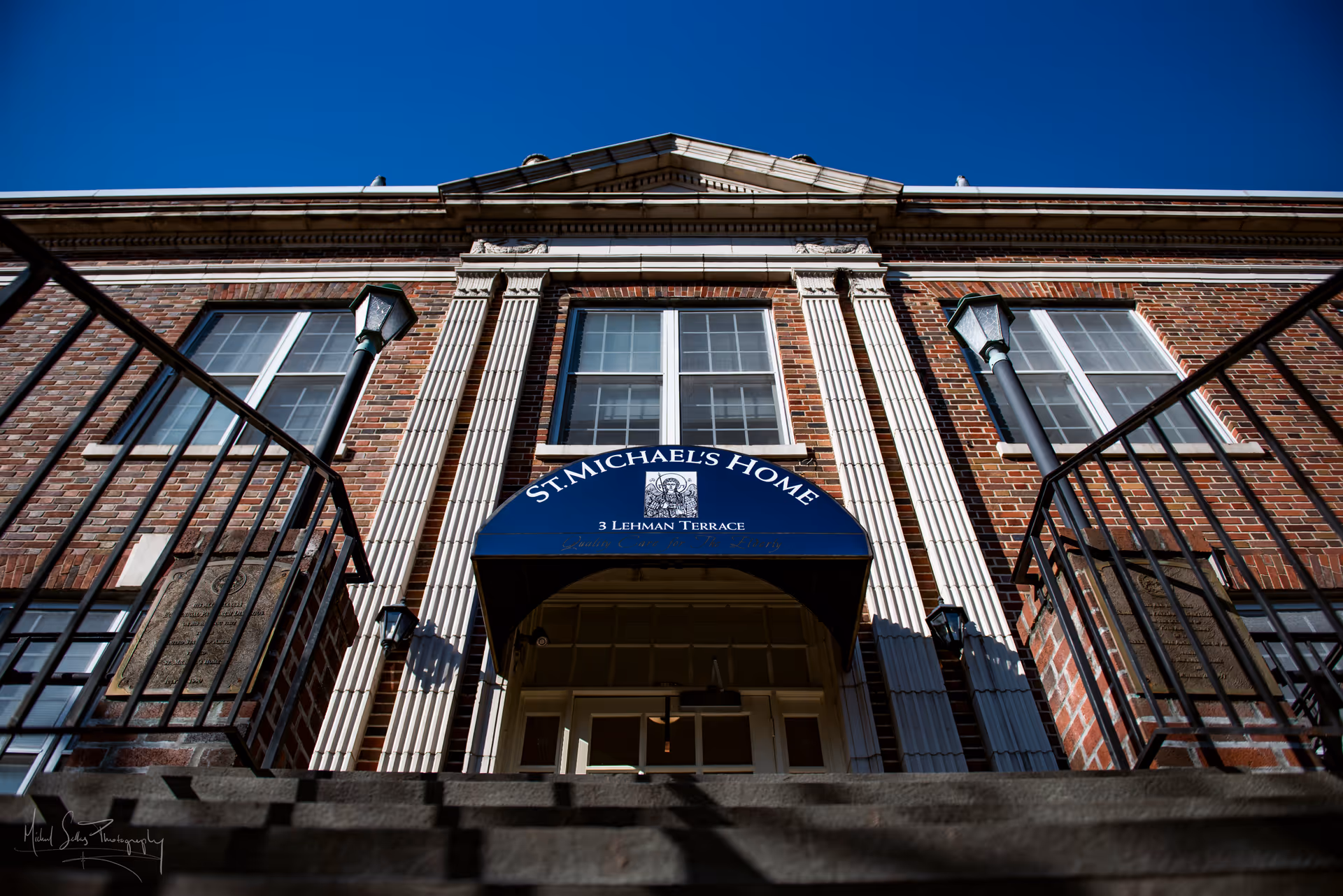 Upward view of the front entrance of St. Michael's Home, a brick building with white columns and a blue awning displaying the facility's name and address at 3 Lehman Terrace. There are black metal railings on both sides of the stairs leading up to the entrance, and two lampposts are visible near the doorway under a clear blue sky.