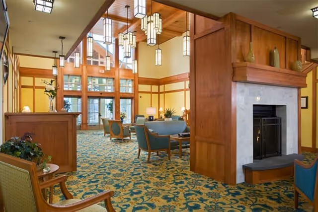 Spacious senior living facility common area with high ceilings, large windows, wooden beams, and a fireplace. The room is furnished with comfortable chairs and sofas arranged around tables, with decorative plants and pendant lighting hanging from the ceiling.