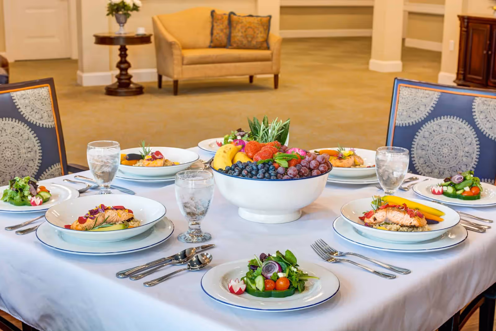 A dining table set with plates of salmon, fresh salads, glasses of water, and a large bowl of assorted fresh fruits including bananas, strawberries, blueberries, and grapes. The table is covered with a white tablecloth and surrounded by chairs with patterned upholstery. In the background, there is a beige loveseat and a small round wooden table with a decorative item on it.