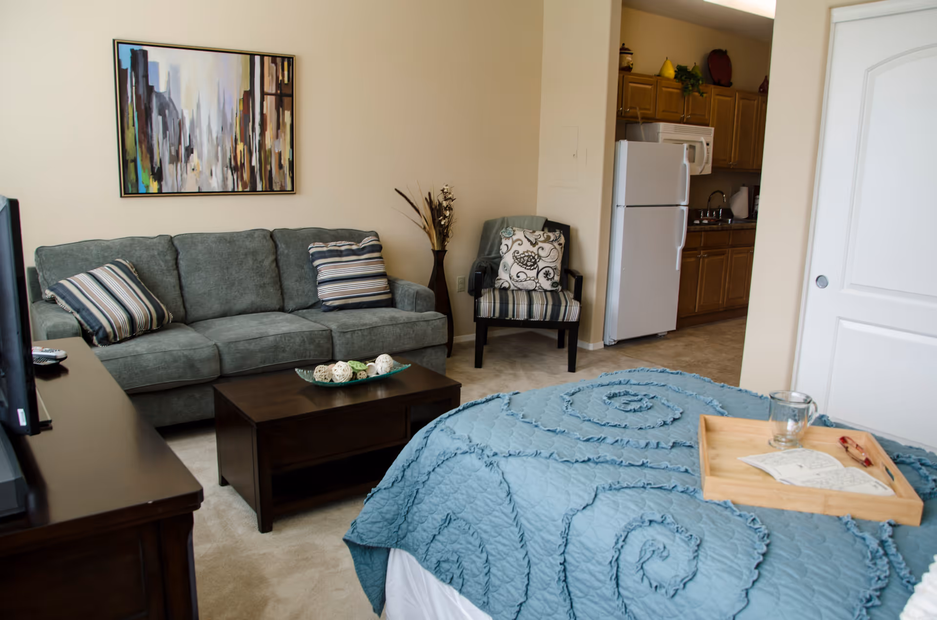 Interior view of a senior living facility room featuring a bed with a blue quilt and a wooden tray holding a glass and a book, a green sofa with striped pillows, a striped armchair, a dark wooden coffee table with decorative items, a TV on a dark wooden stand, and a kitchen area with a refrigerator, microwave, and wooden cabinets in the background.