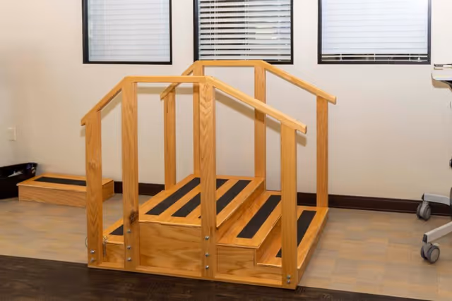 A wooden therapy staircase with handrails on both sides and non-slip black strips on each step, placed indoors in front of three windows with closed blinds.