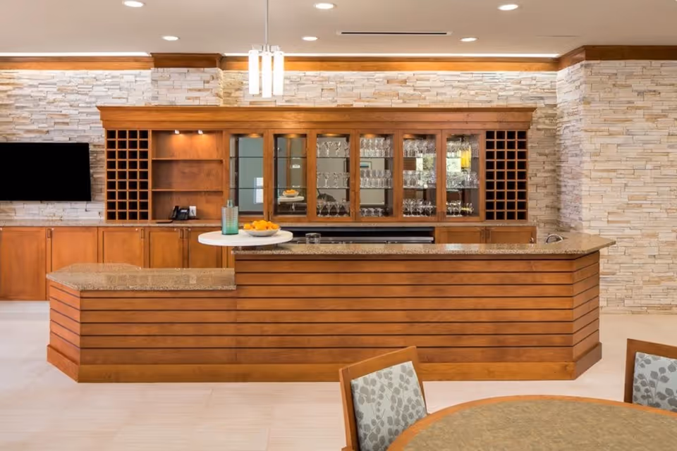 A modern bar area with wooden cabinetry and a granite countertop. The back wall features glass cabinets filled with glassware, wine racks on either side, and a mounted flat-screen TV on the left. A small round table with a glass pitcher and bowl of oranges is placed on the countertop. The room has light-colored stone walls and recessed lighting.