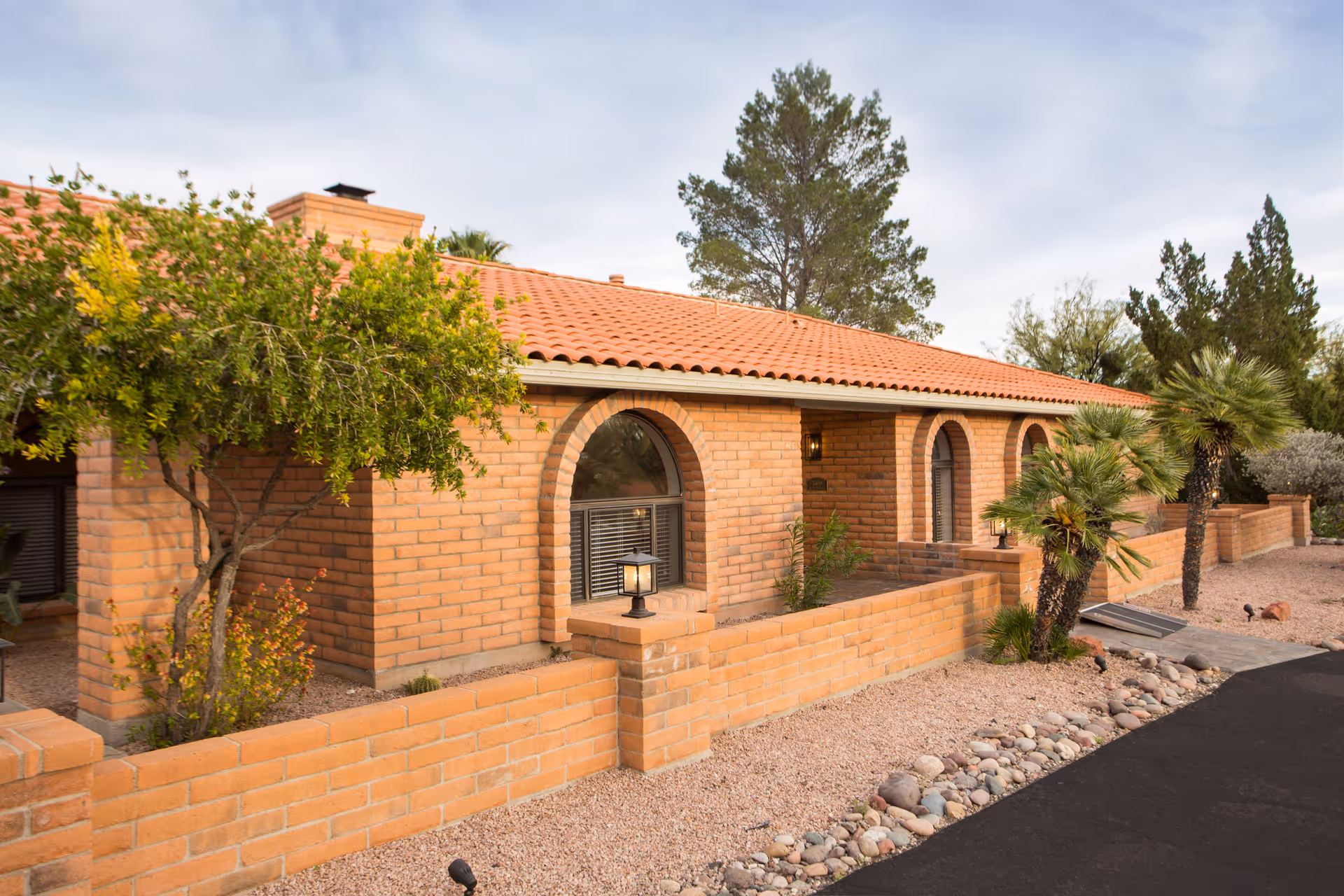 Exterior view of a single-story brick building with a red tile roof, arched windows, and a small enclosed front patio area with desert landscaping including small palm trees and gravel ground cover.