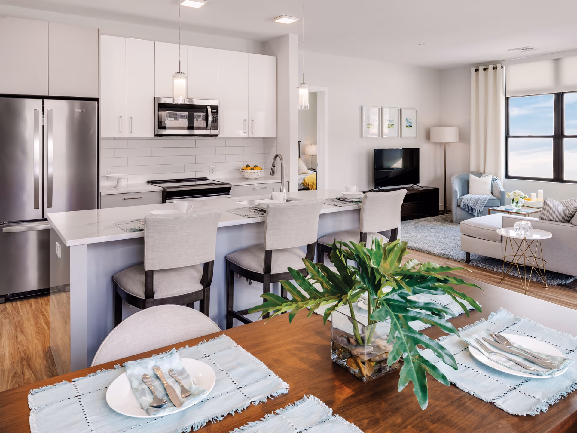 A modern open-concept living space featuring a kitchen with white cabinets, stainless steel refrigerator, microwave, and stove. A kitchen island with three upholstered bar stools is set with placemats and cups. Adjacent to the kitchen is a living room area with a TV on a stand, a blue armchair, a floor lamp, and a large window with white curtains. In the foreground, a wooden dining table is set with plates, silverware, light blue placemats, and a glass vase with green leafy plants.