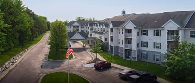 Exterior view of a multi-story senior living facility named Lockwood of Genesee, showing a driveway with parked cars, a covered entrance, an American flag, and surrounding greenery.