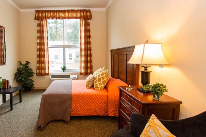 A cozy bedroom with a bed covered in an orange quilt and brown blanket, two decorative pillows, a wooden headboard, a wooden nightstand with a lamp and a small plant, a window with plaid curtains, a small table with a plant, and a cushioned chair with a patterned pillow.