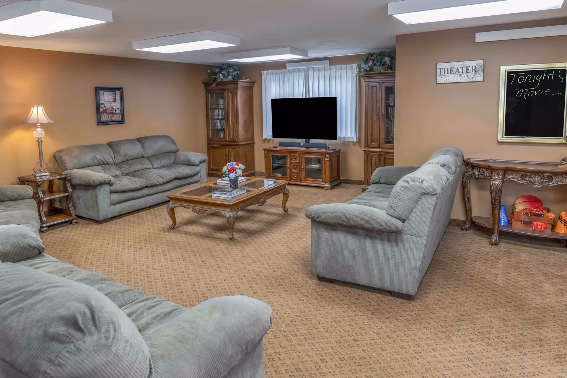 A cozy living room with three green upholstered sofas arranged around a wooden coffee table with books and a flower vase. A flat-screen TV is centered on a wooden TV stand between two wooden display cabinets. The walls are painted beige, and there is a lamp on a side table to the left. A sign on the wall reads 'THEATER' and a chalkboard says 'Tonight's movie...'. The carpet is patterned in a light brown color.