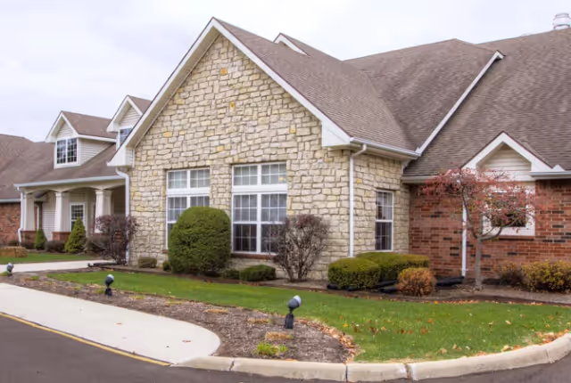 Exterior view of a senior living facility building with stone and brick facade, multiple windows, a sloped roof, and landscaped bushes and trees along a curved driveway.