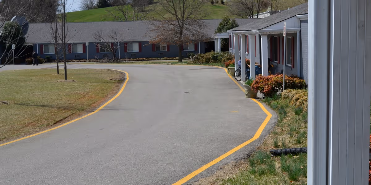 View of the exterior driveway and entrance area of North Roanoke Assisted Living facility, showing a paved road with yellow lines, a grassy area with young trees, and a single-story building with a covered porch where a person in a wheelchair is seated.