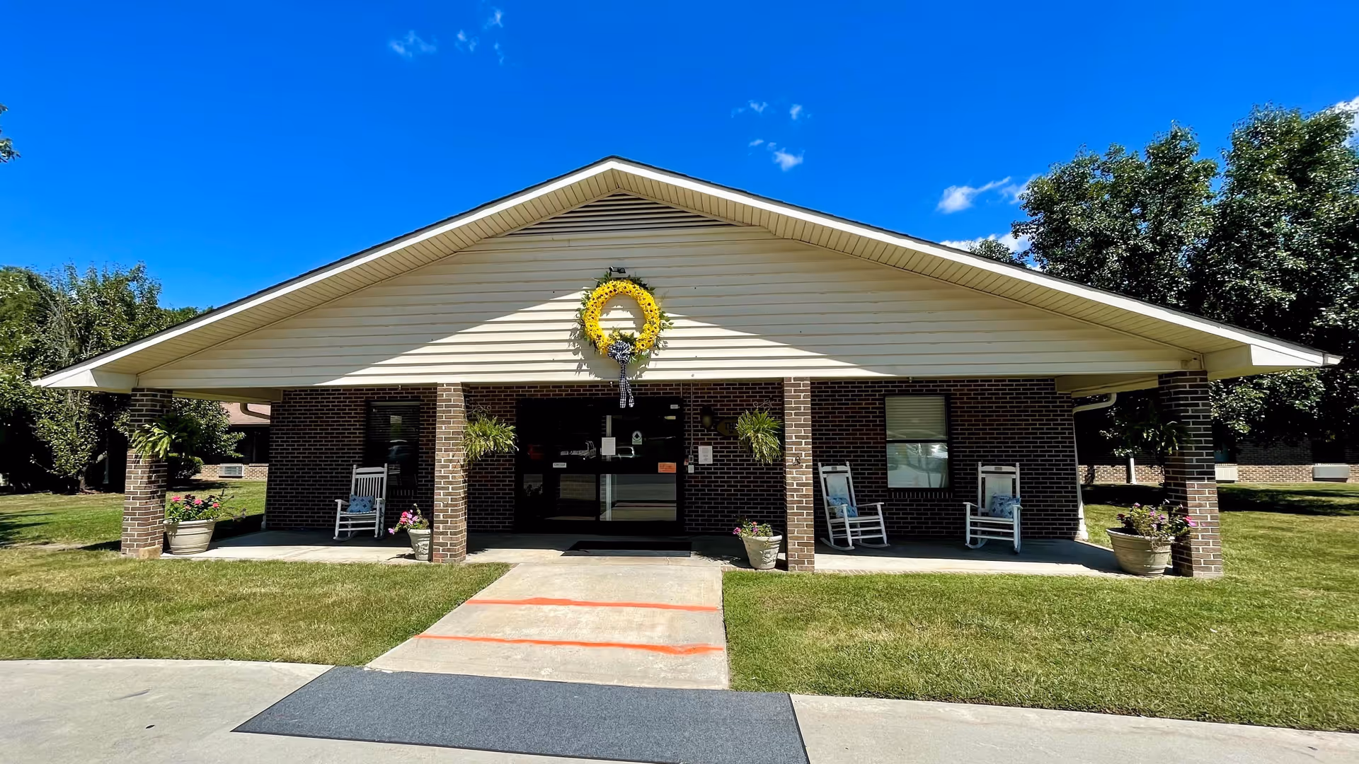 Front exterior view of a single-story brick building with a peaked roof and a large yellow wreath hanging above the entrance. The entrance is covered by a porch supported by brick columns, with rocking chairs and potted plants on either side. The building is surrounded by green grass and trees under a clear blue sky.