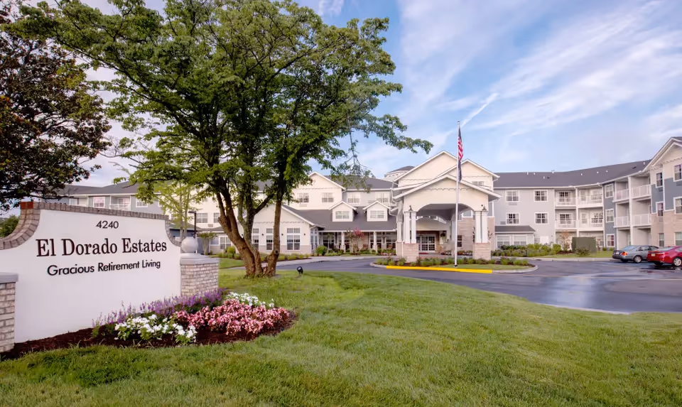 Exterior view of El Dorado Estates Gracious Retirement Living facility with a large sign displaying the name and address 4240. The building is a multi-story structure with balconies, surrounded by well-maintained green lawns, trees, and flower beds. There is a driveway with parked cars and an American flag flying on a flagpole near the entrance.
