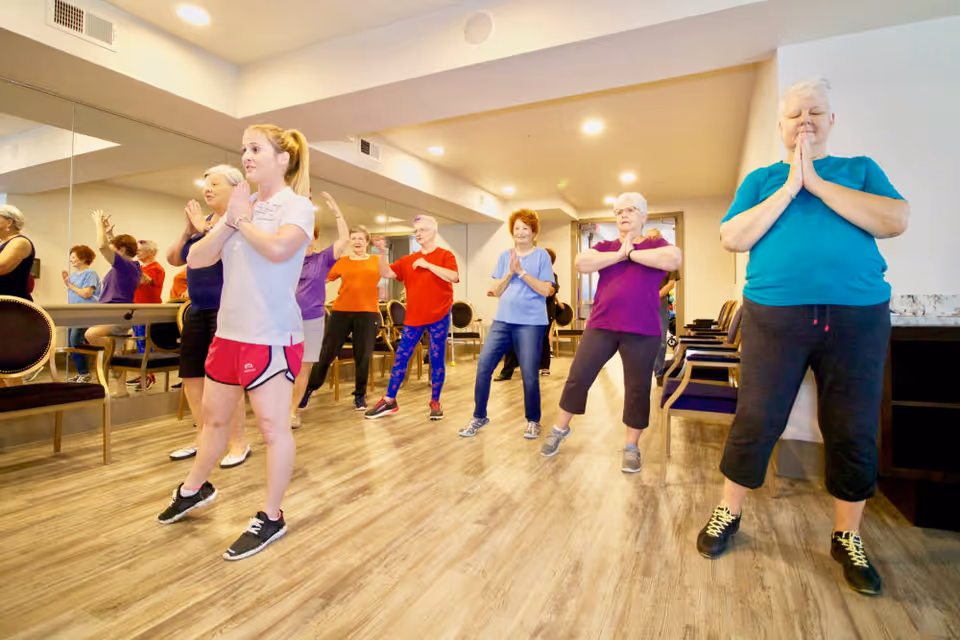 A group of senior women and a younger instructor participating in a fitness or yoga class inside a well-lit room with wooden flooring and mirrored walls. The participants are standing spaced apart, some with hands pressed together in a prayer position, engaging in a stretching or balance exercise.