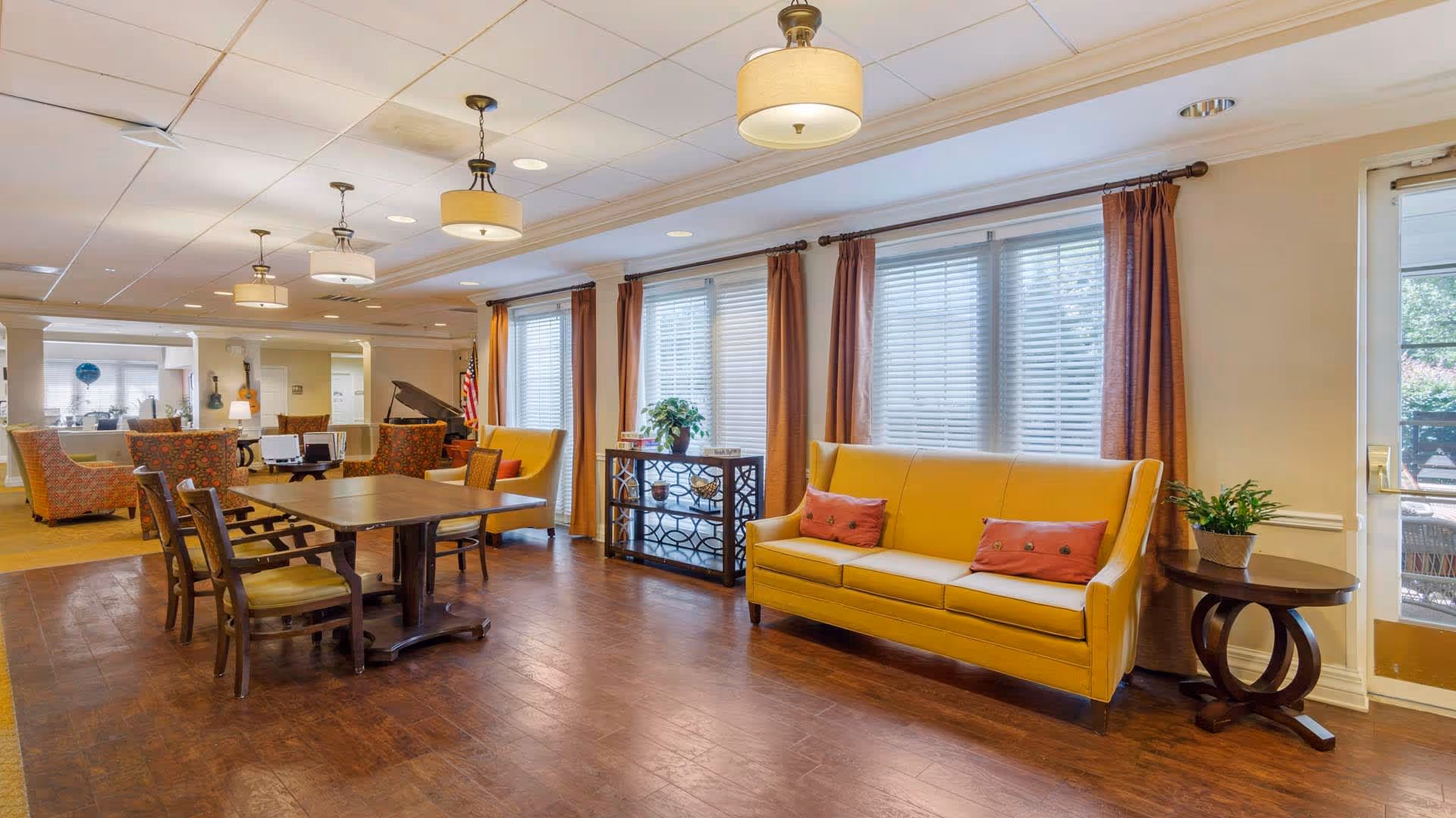A spacious and well-lit living room area in a senior living facility featuring a yellow sofa with red cushions, a wooden table with chairs, large windows with blinds and brown curtains, and decorative plants on side tables. The room has wooden flooring and ceiling lights, with additional seating and a piano visible in the background.