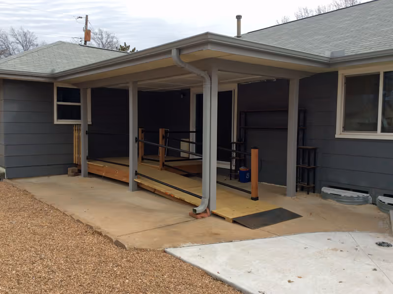 Exterior view of a gray building with a covered porch area featuring a wooden wheelchair ramp with handrails. The ramp leads to a door under the porch roof. The ground around the porch is a mix of concrete and gravel.