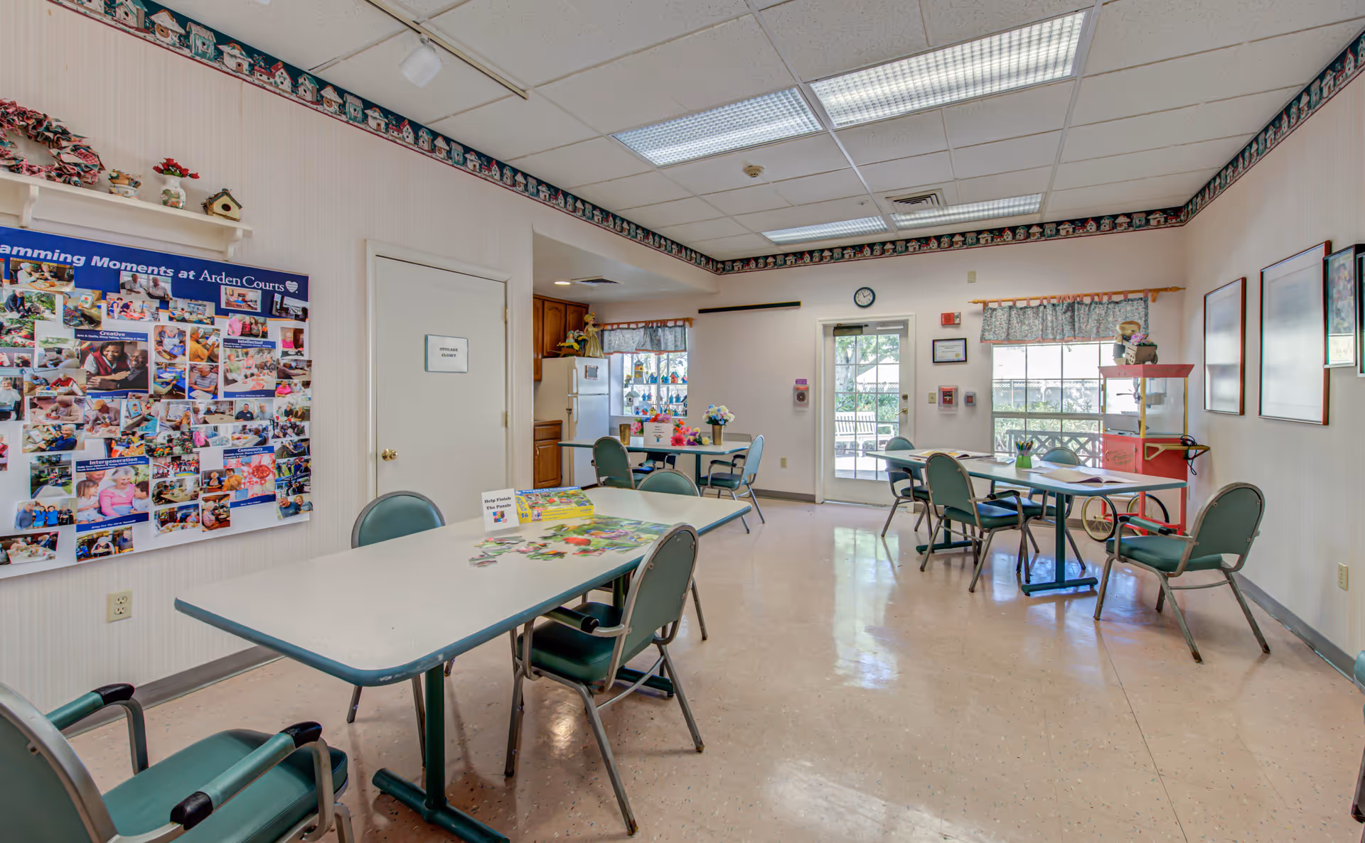 A bright activity room in a senior living facility with several tables and green chairs arranged around them. On one table, there is a partially completed puzzle and a sign that says 'Help Finish The Puzzle.' The walls are decorated with a bulletin board displaying photos titled 'Programming Moments at Arden Courts,' a popcorn machine, framed pictures, and a floral border near the ceiling. There is a door labeled 'Storage Closet,' a small kitchen area with a refrigerator, and windows with floral curtains letting in natural light.