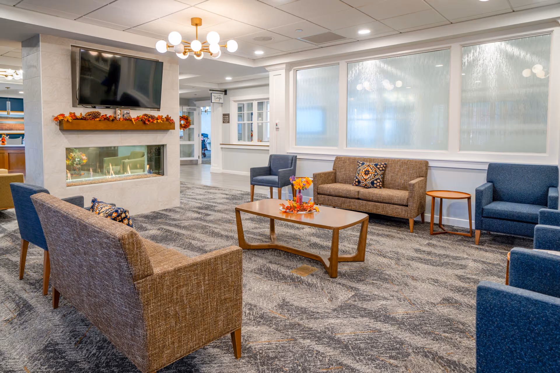 A cozy senior living facility common area with a modern fireplace, a wall-mounted TV above it, and comfortable seating including brown and blue armchairs and sofas arranged around a wooden coffee table decorated with a fall-themed centerpiece. The room has patterned carpet flooring, white walls with large frosted windows, and a ceiling light fixture.