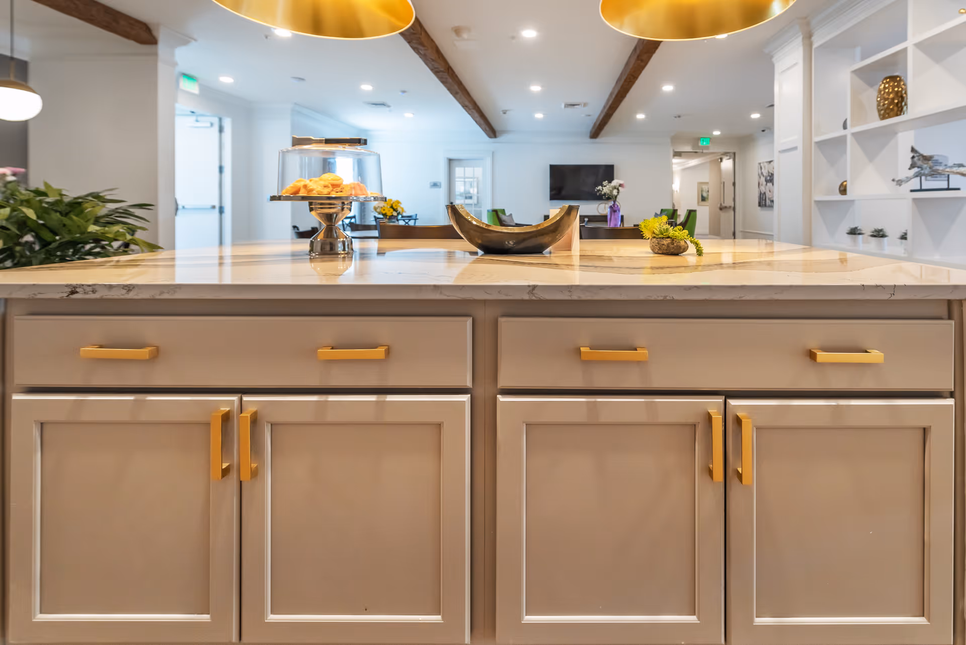 View of a modern kitchen island with beige cabinets and gold handles, topped with a marble countertop. On the countertop, there is a glass cake stand with pastries, a decorative bowl, and a small plant. In the background, a living area with a TV mounted on the wall, chairs, and decorative shelves is visible.