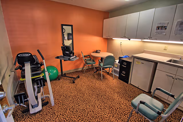 A small room with exercise equipment including a stationary bike and a green exercise ball, a round table with three chairs, a wall-mounted mirror, white cabinets, a small refrigerator, a sink, and a filing cabinet. The walls are painted orange and beige, and the floor is covered with patterned carpet.
