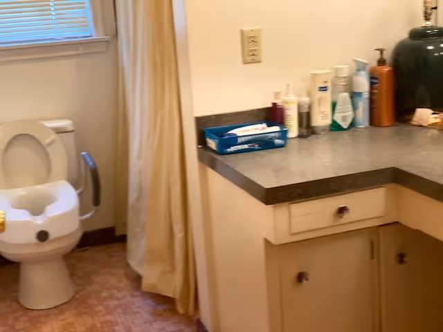 A bathroom with a toilet equipped with armrests and a raised seat, a window with blinds, a shower curtain, and a countertop with various toiletries and hygiene products.