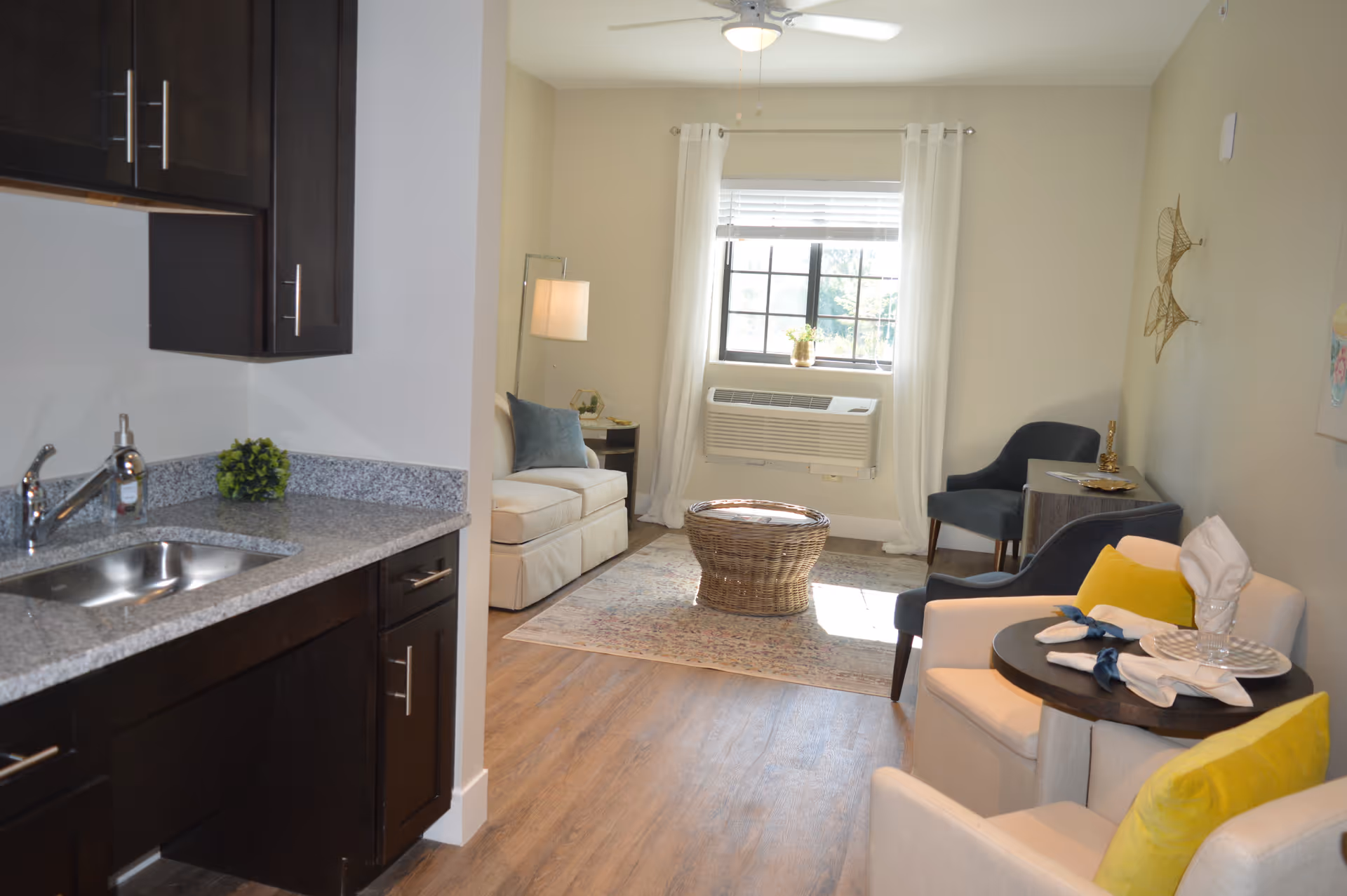 A cozy living area with a small kitchenette on the left featuring dark cabinets and a granite countertop with a sink. The living space includes a beige sofa, a round wicker coffee table on a patterned rug, two dark blue chairs, and two beige armchairs with yellow pillows and a small round table set with plates and napkins. A window with white curtains and an air conditioning unit is in the background, along with a ceiling fan above.